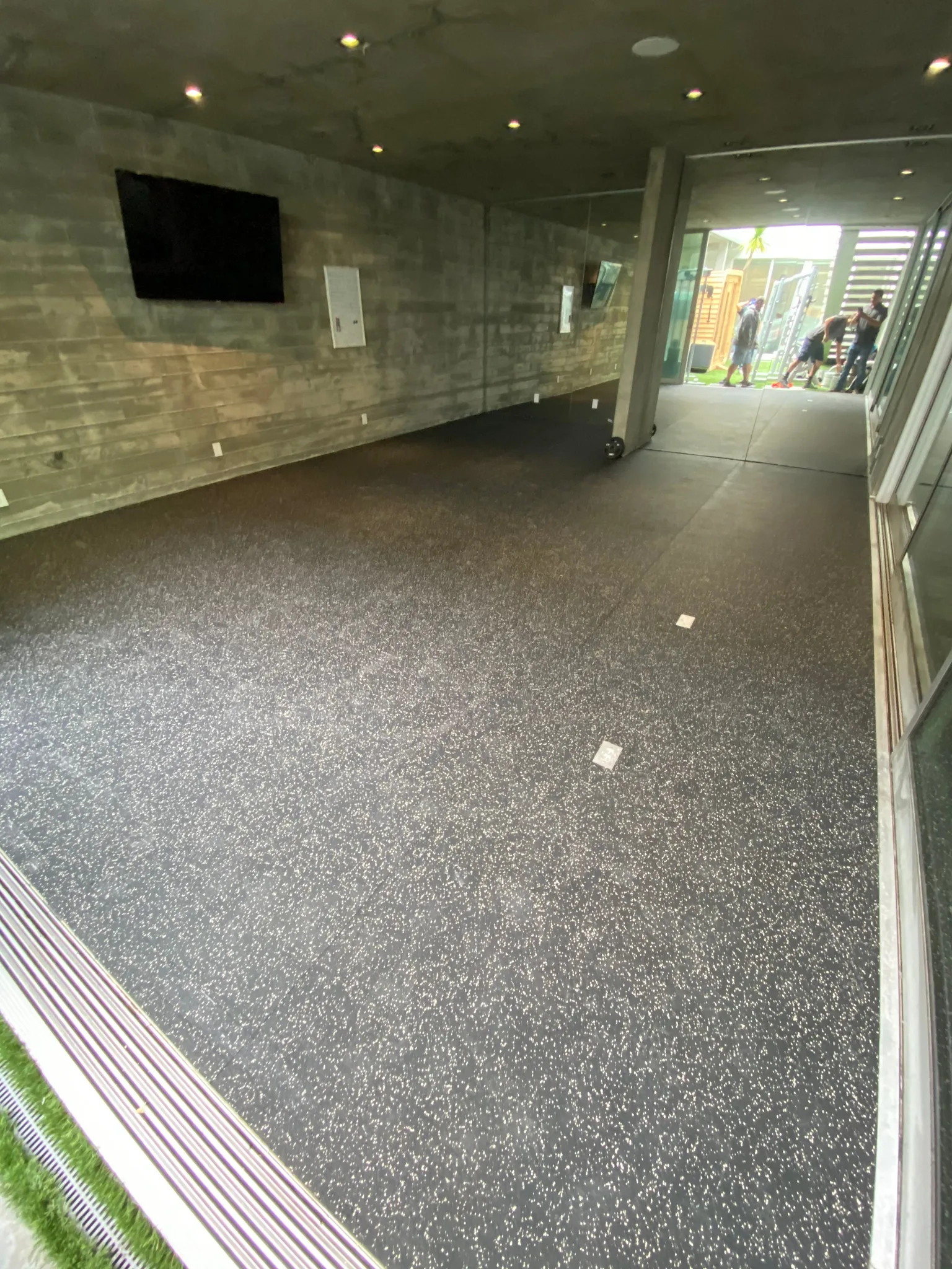 Speckled black rubber flooring in a board-formed concrete gym opening onto a garden