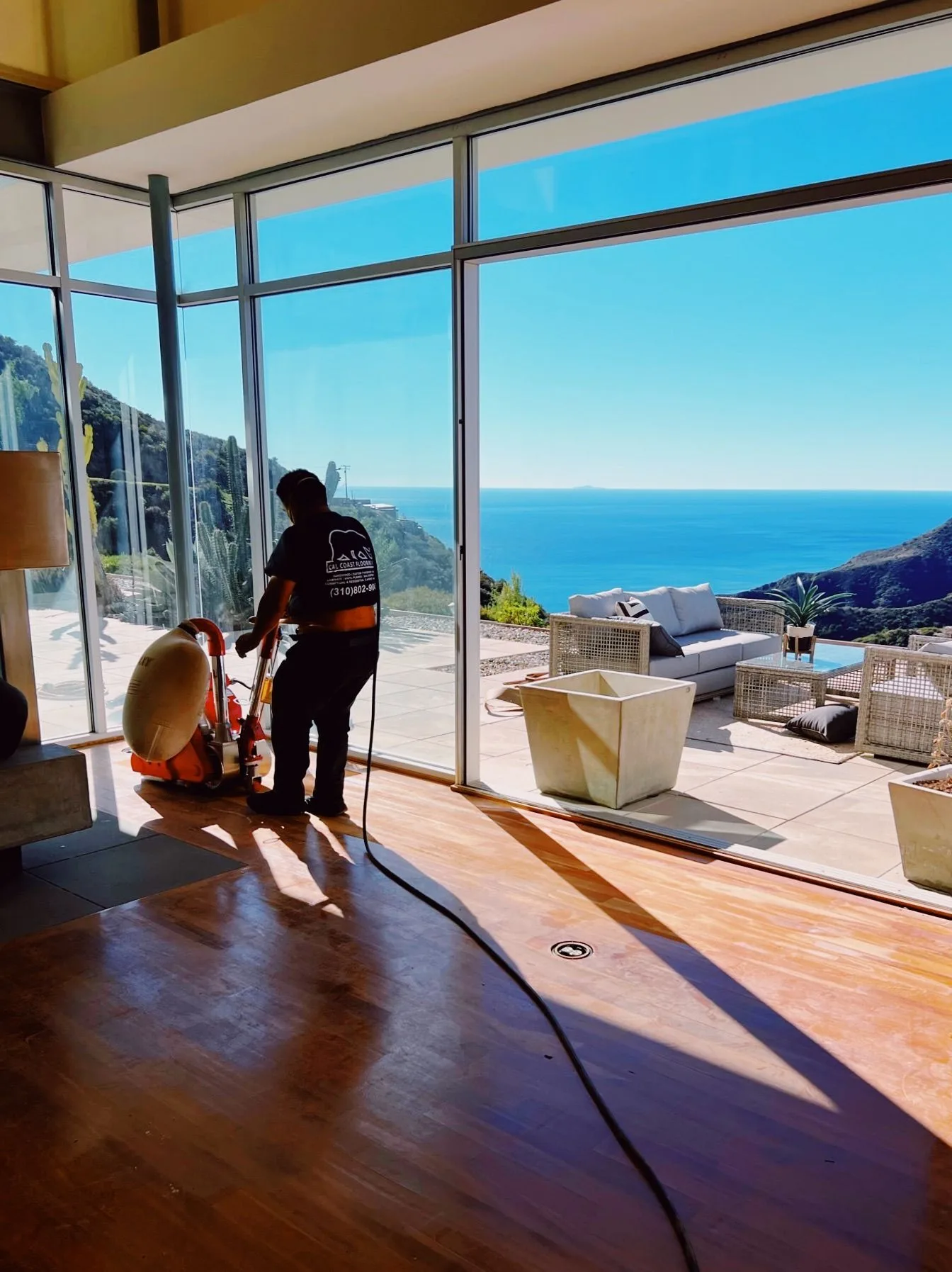 Sanding a dark hardwood floor in a glass-walled room above the coast