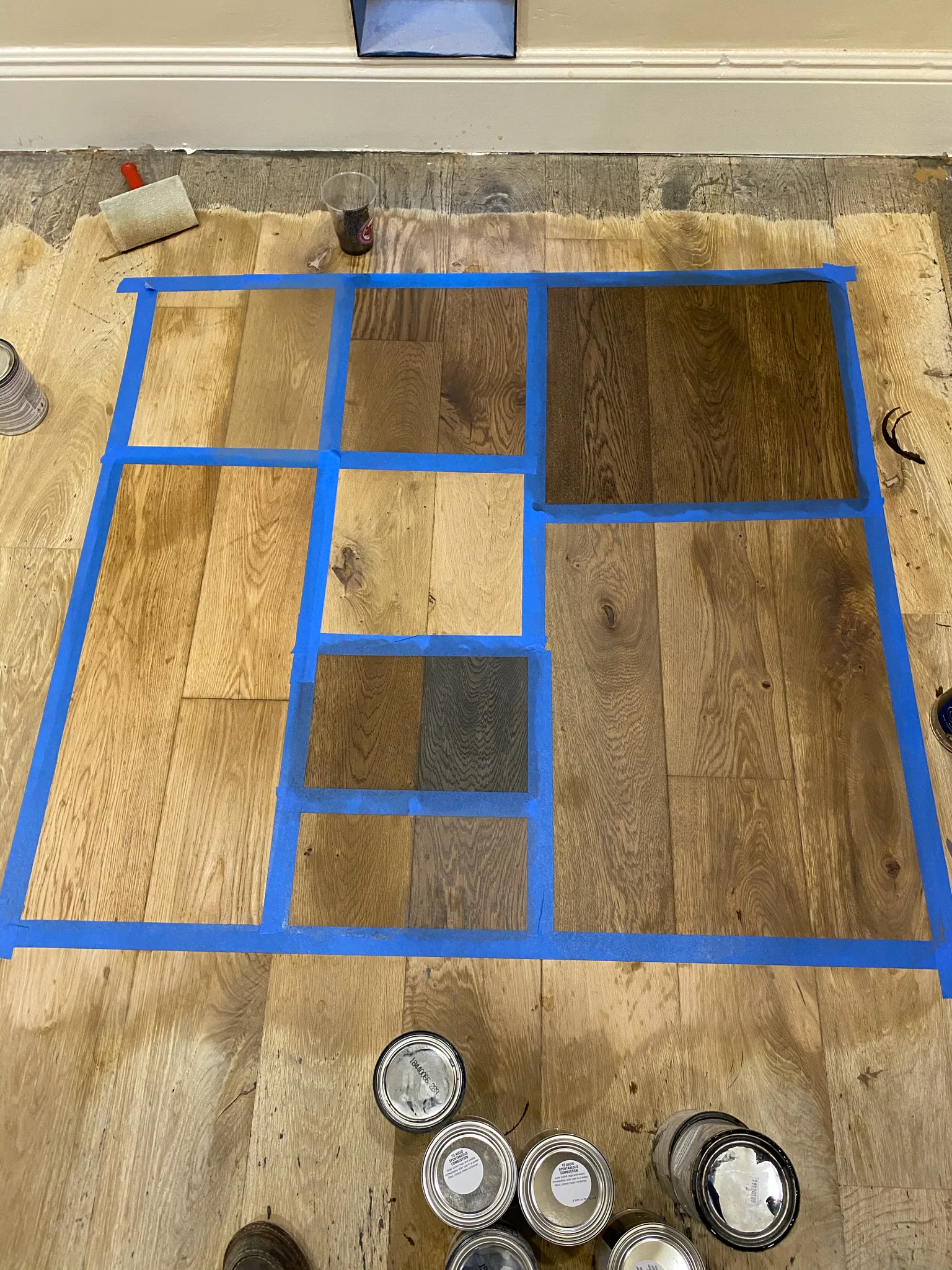 Taped-off grid of custom stain samples on the sanded hardwood floor at Nando Trattoria, with the stain cans lined up beside it